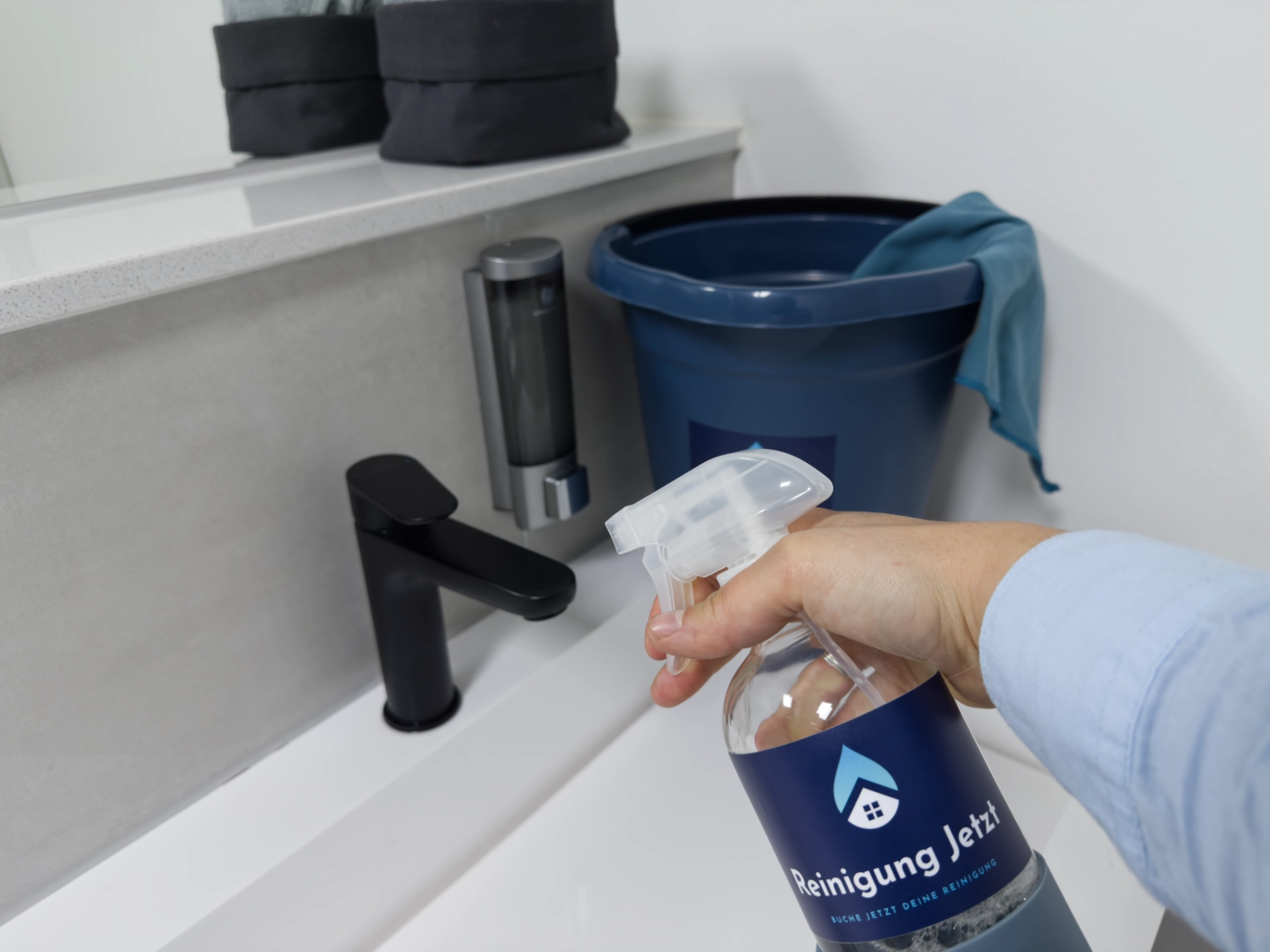 Eine Person reinigt ein modernes Badezimmerwaschbecken mit einer Sprühflasche, die das Logo 'Reinigung Jetzt' trägt. Im Hintergrund steht ein blauer Putzeimer mit einem Tuch. Das Waschbecken hat einen schwarzen Wasserhahn und eine Seifenspender-Wandhalterung.